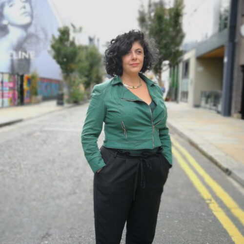 A woman standing in the street with her hands in her pockets, wearing a green top.