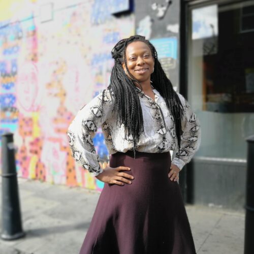 Vicki Dela Amedume stands in a street with her hands on her hips, smiling. She is a Black African Female in her forties with long black braids