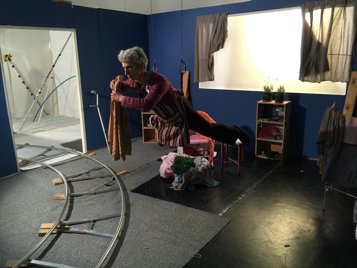An elderly woman floats in the air using a harness, in a set that looks like a house.