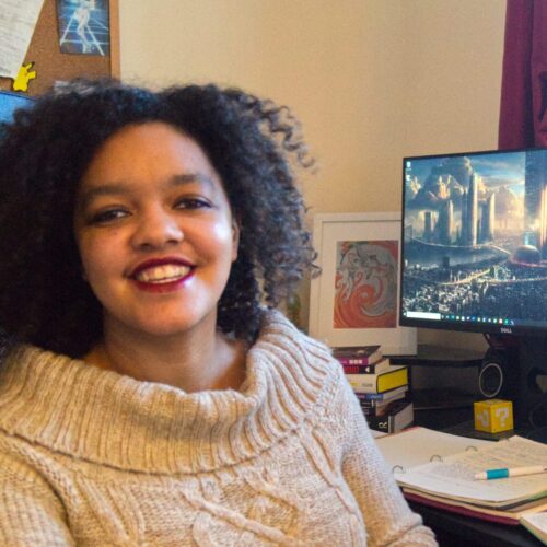 A mixed race woman sits in front of two computer screens, smiling.