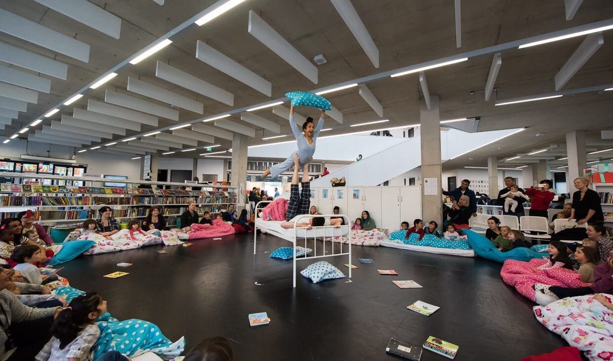 An acrobat balances on another's feet while holding onto a pillow. They are in a library space with an audience of children watching