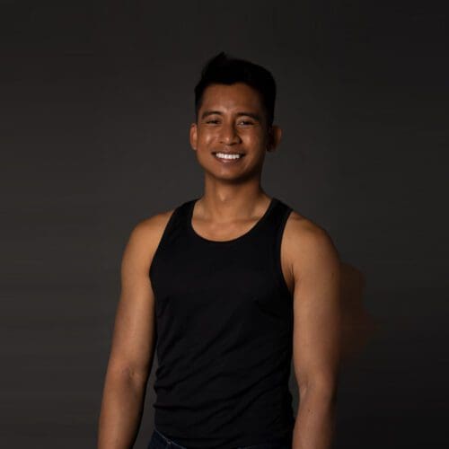 A singaporean-born male with short black hair and black tank top smiles in front of a dark grey backdrop