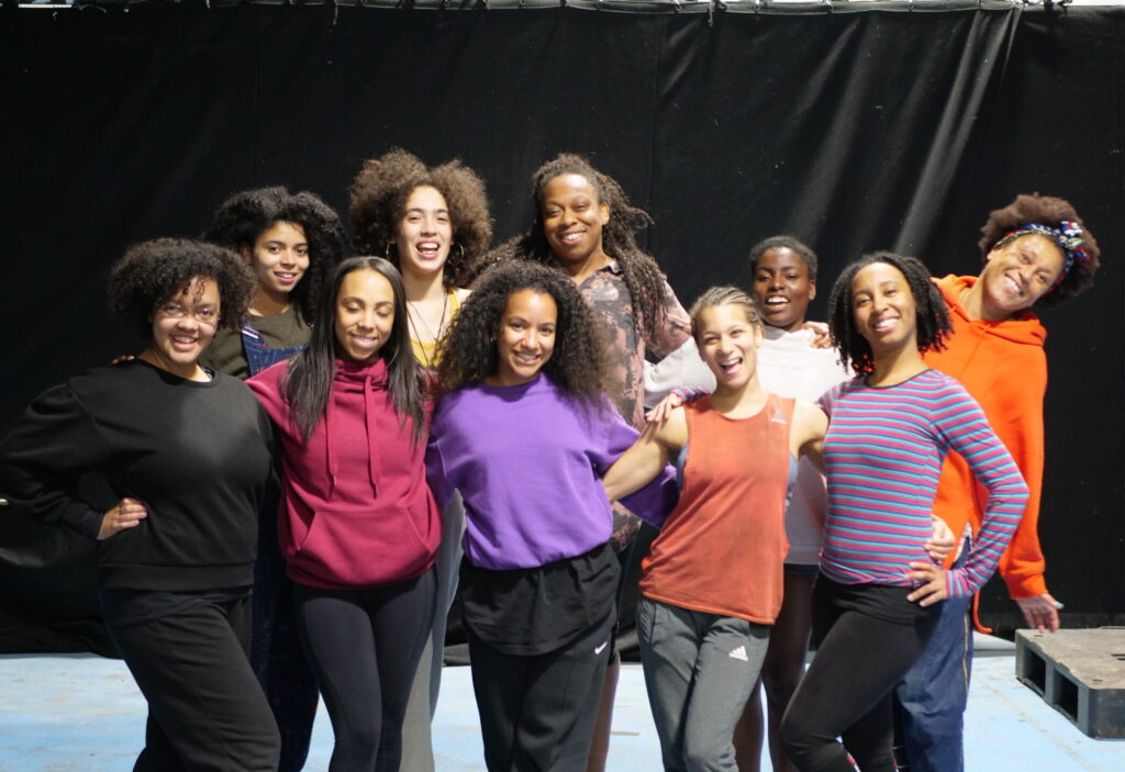 A group of 12 black female and non-binary circus artists stand together in a rehearsal room smiling