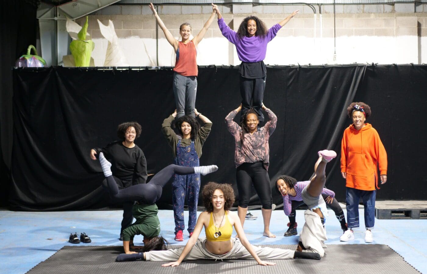 A group of Black female and non-binary circus artists stand together in a rehearsal room. some are standing on each others shoulders creating a pyramid with their arms outstretched wide.