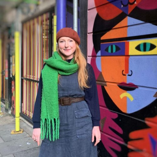 A white woman in her thirties with long ginger hair, wearing a denim dress, orange beret and green scarf stands in front of a graffiti wall