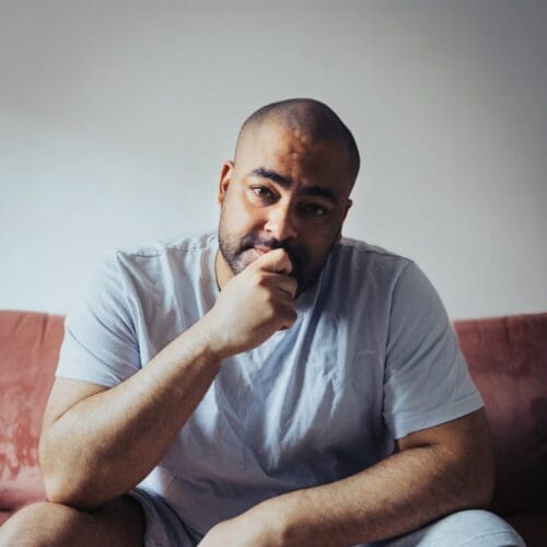 A man sits down with his chin resting in his hand. He has short black hair and a beard. He wears a white tshirt