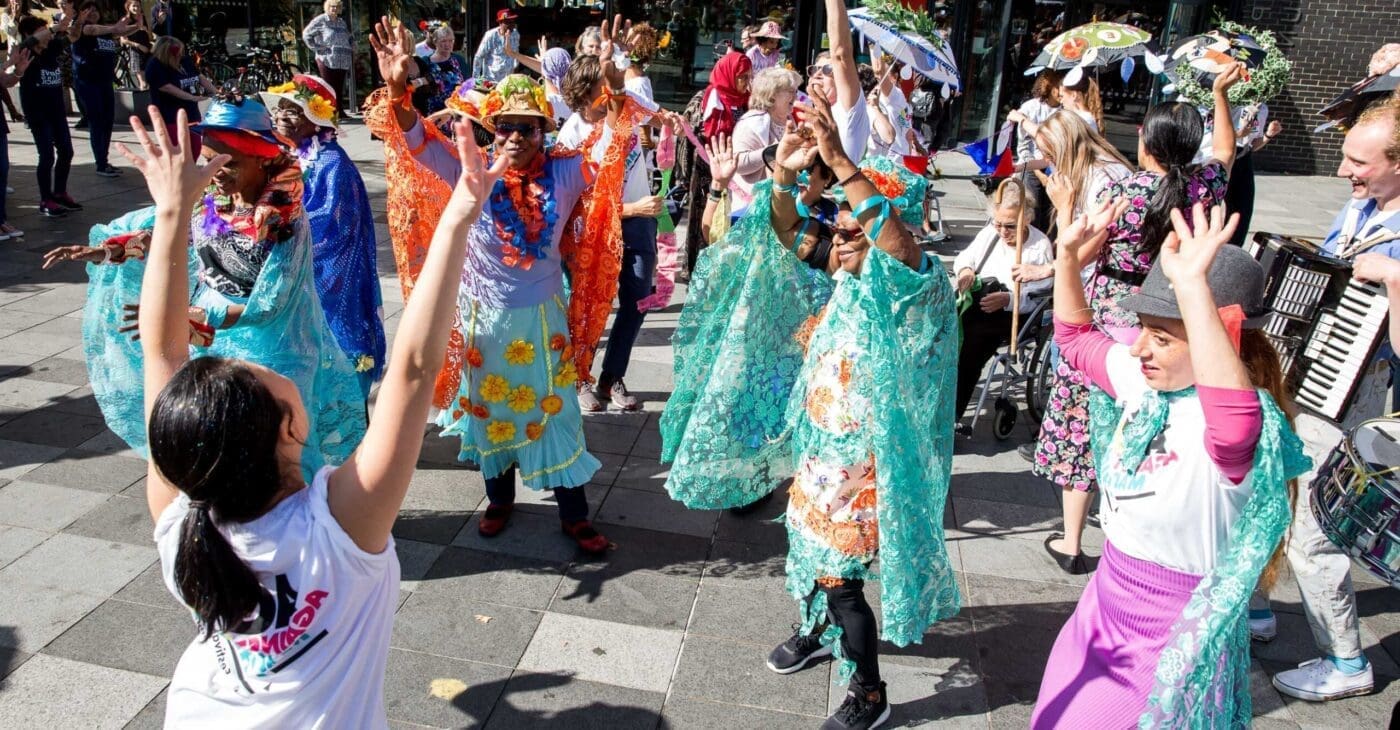 A big group of people throw their hands up in dance wearing brightly coloured carnival costumes - led by charlene low our workshop leader