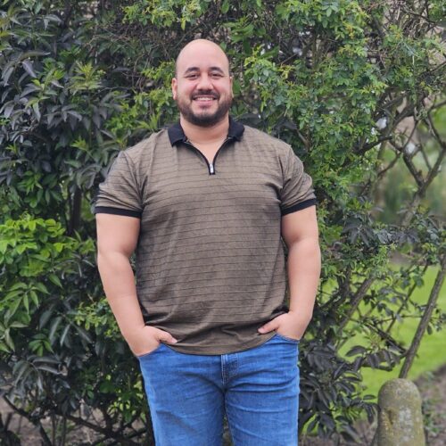 a 34 year old mixed race man stands smiling with his hands in his pockets in front of some bushes