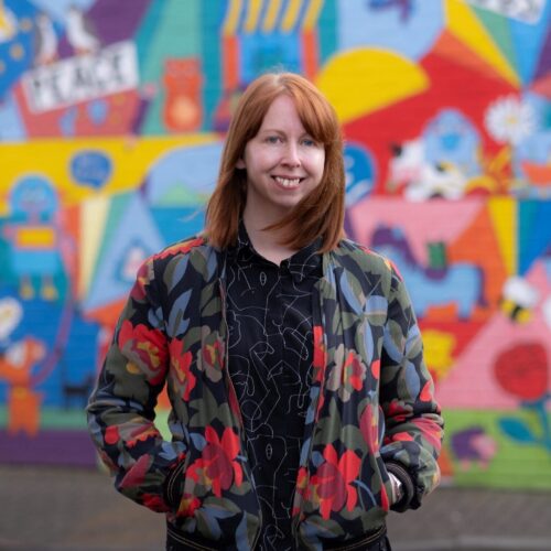 A white woman with ginger hair in her thirties wears a bomber jacket and stands in front of a colourful graffiti wall outside