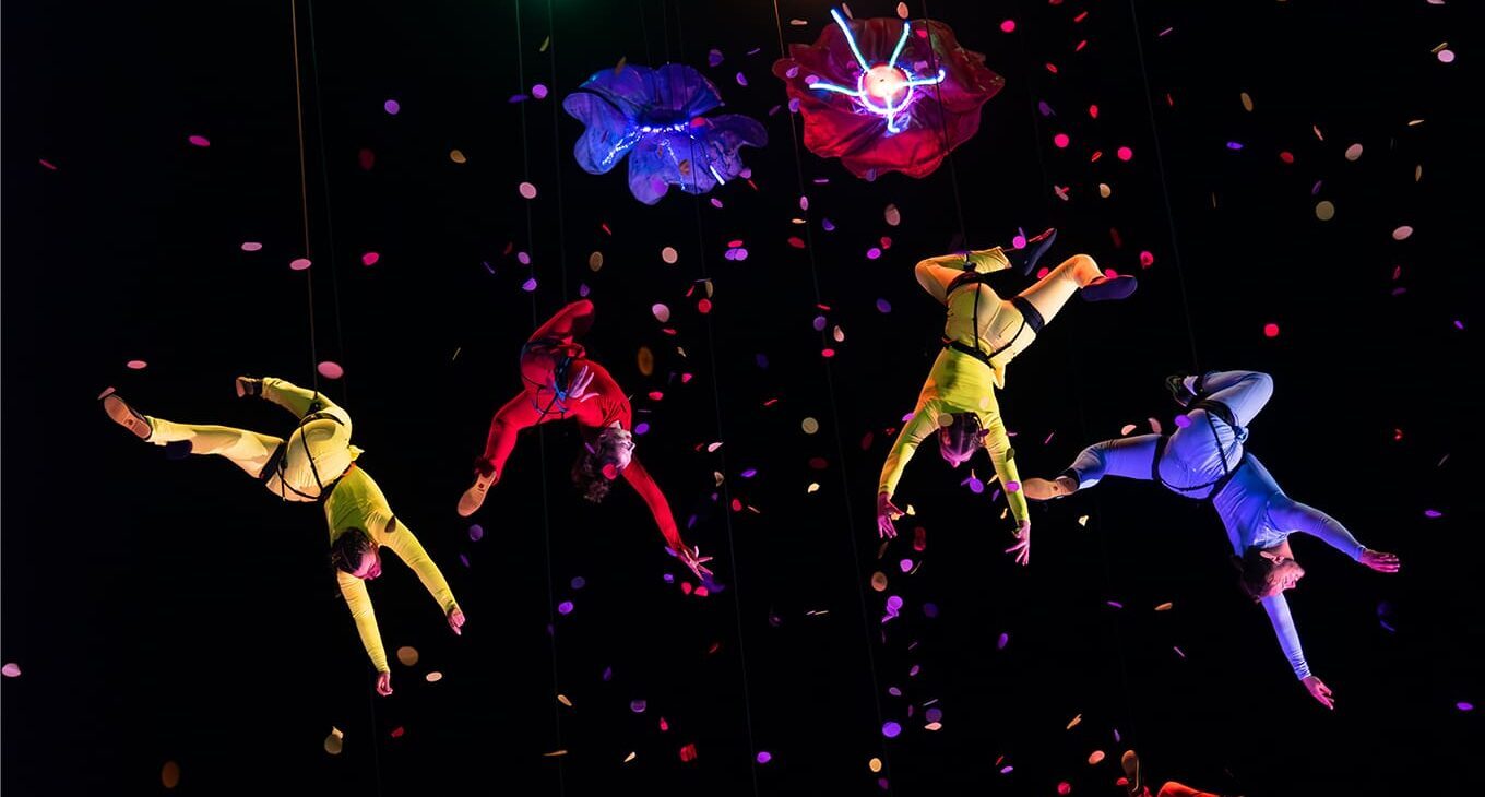 A group of acrobats and aerialists are dangling in the night sky from a huge flower-shaped structure with confetti all around them.