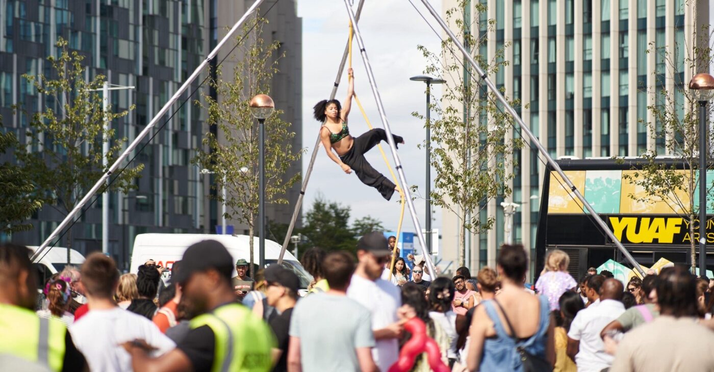 A black female aerialist dangles above a crowd on an aerial rope and outdoor aerial rig. She is in front of large city buildings. There is a diverse crowd all around her